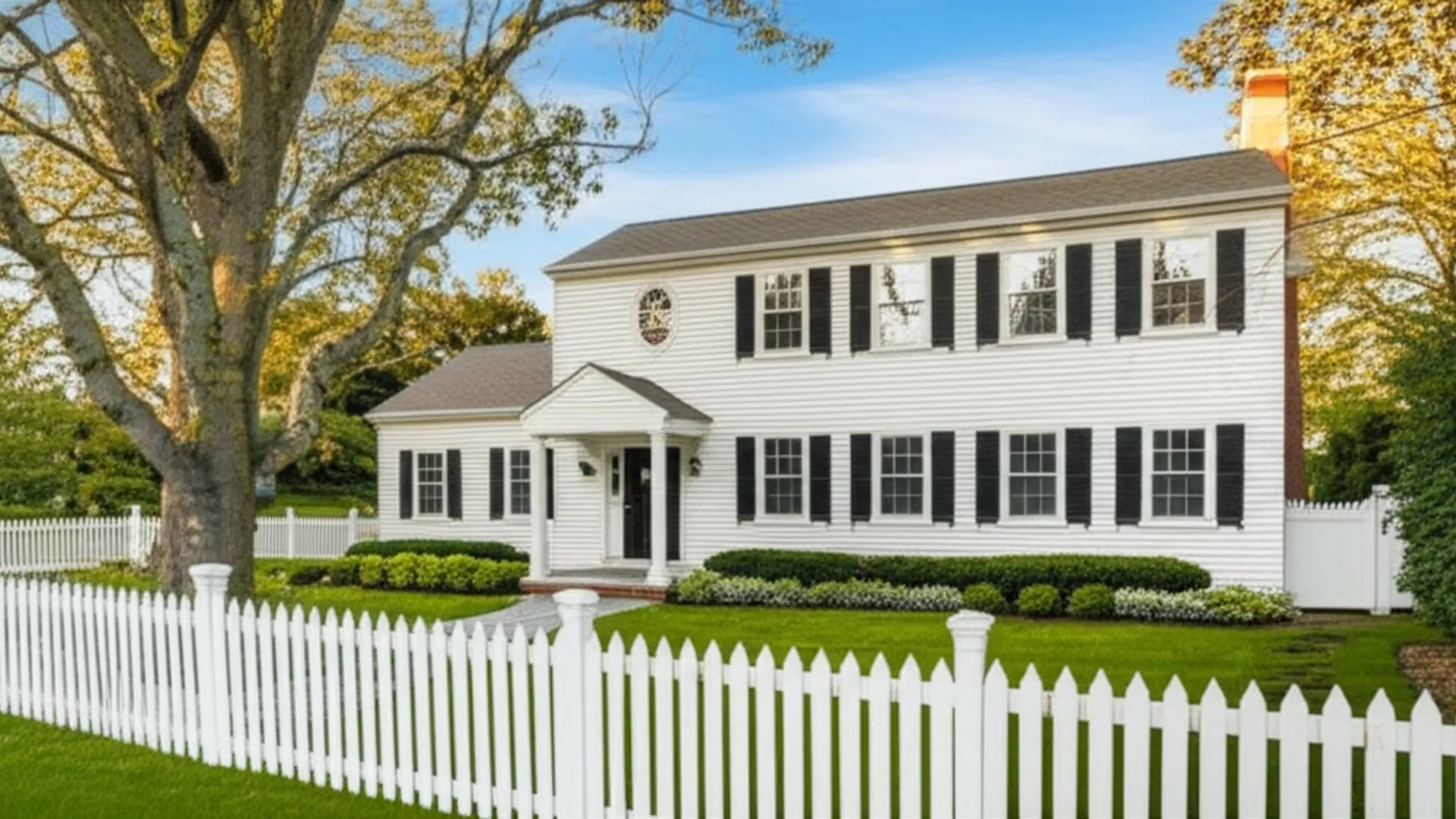 A white clapboard colonial house with black shutters behind a white picket fence, a mature tree beside it in autumn light
